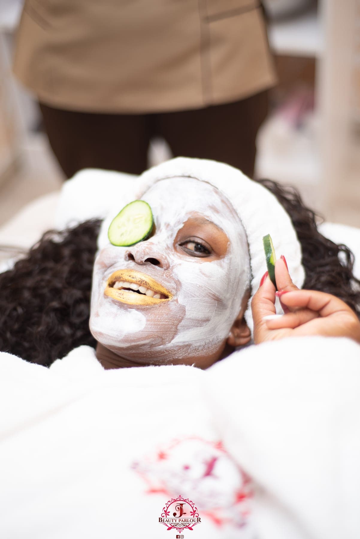Woman smiling under a white face mask with cucumber slices at a spa