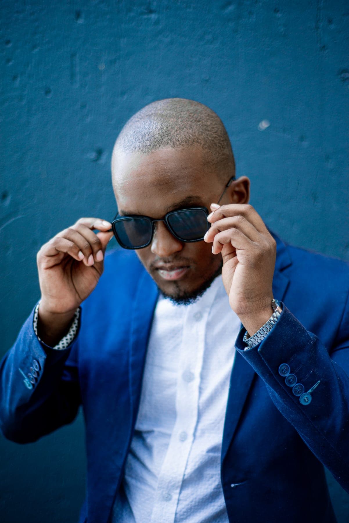 Lowering his sunglasses in a blue blazer against a blue wall