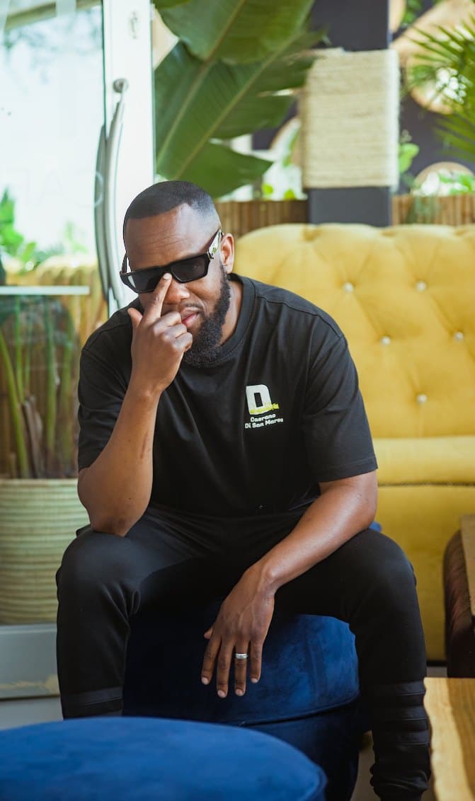 Seated in black, adjusting dark glasses beside a yellow couch