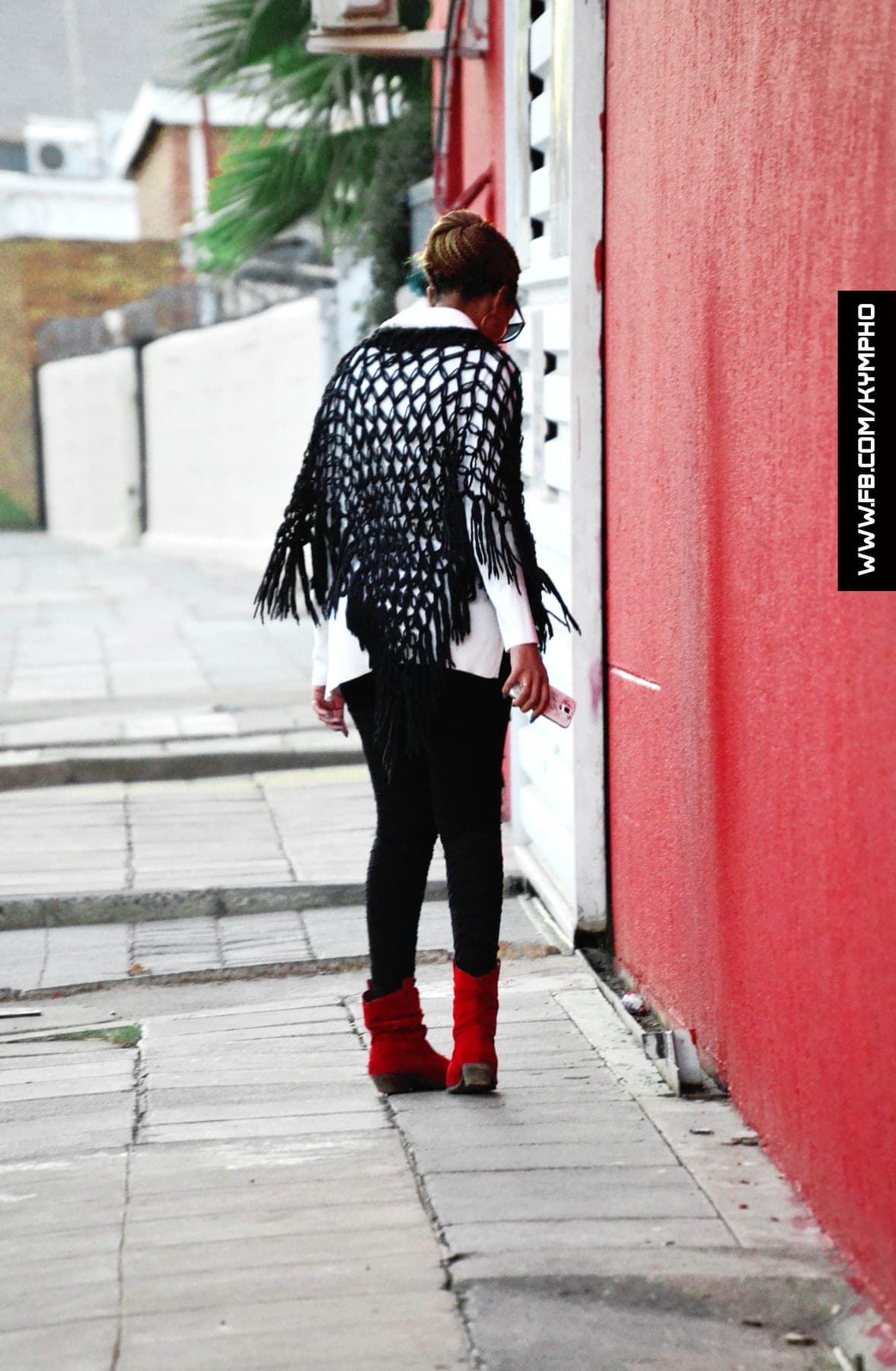 Walking past a red wall in a black net shawl and red boots, phone in hand