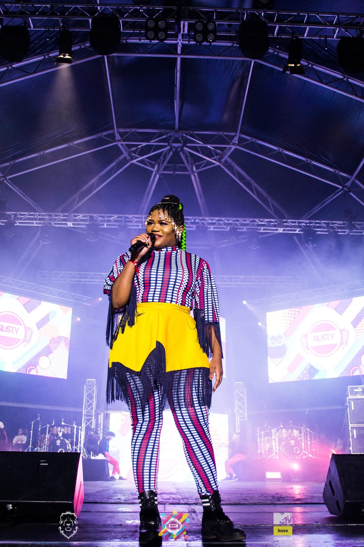Bucie singing in a striped dress and yellow skirt under a purple rig