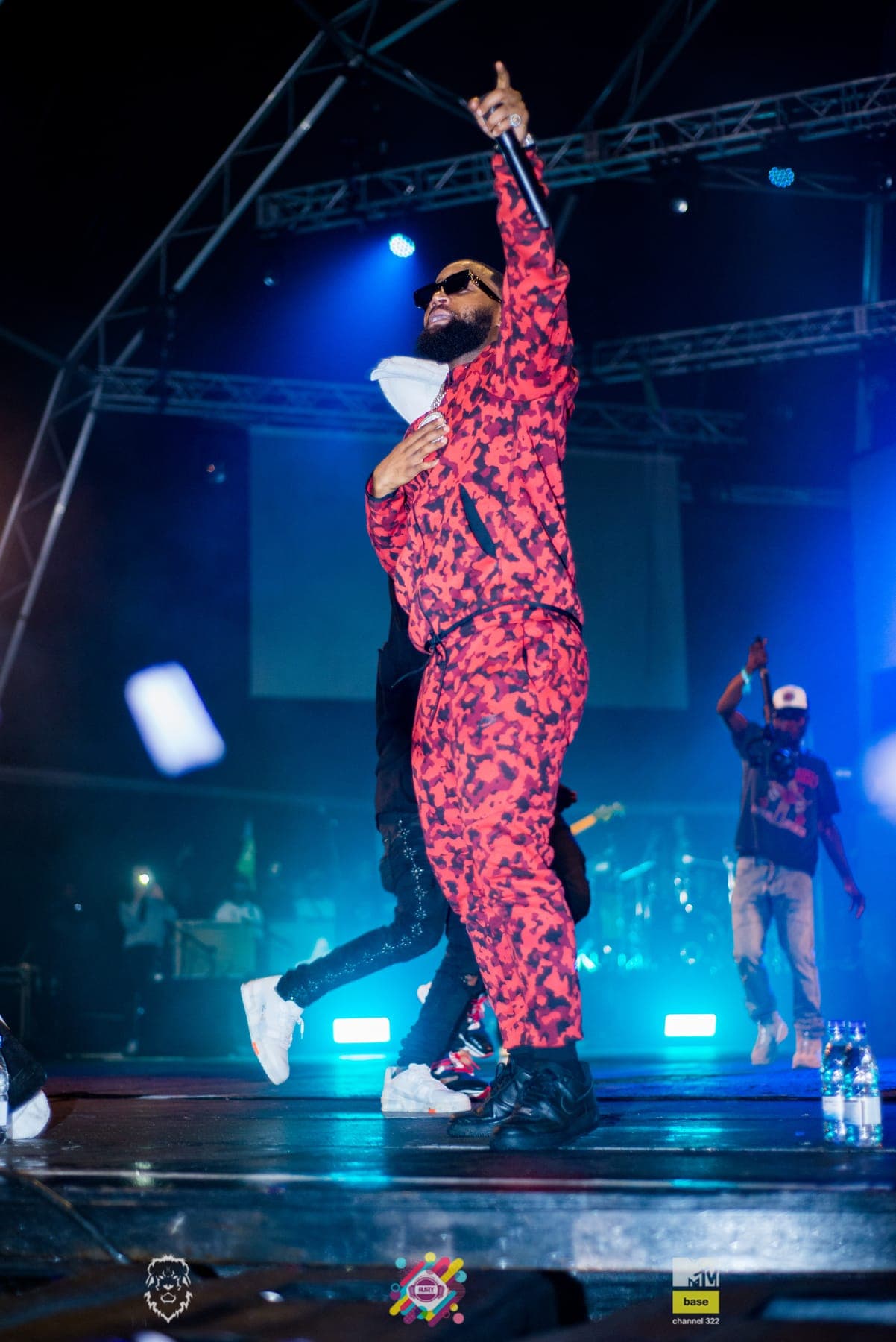 Cassper Nyovest in a red camo tracksuit pointing skyward under blue light