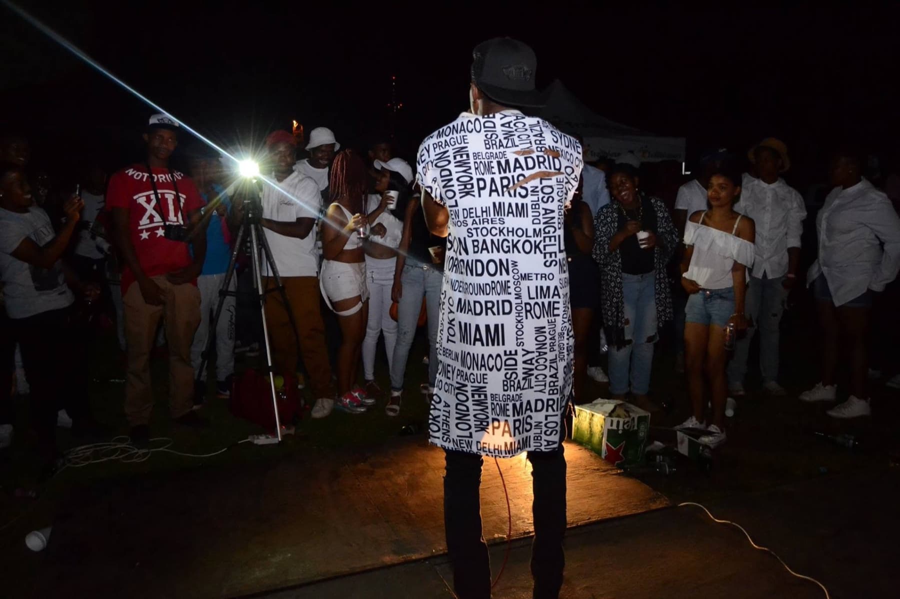 Performer in a city-names shirt facing the crowd under one spotlight