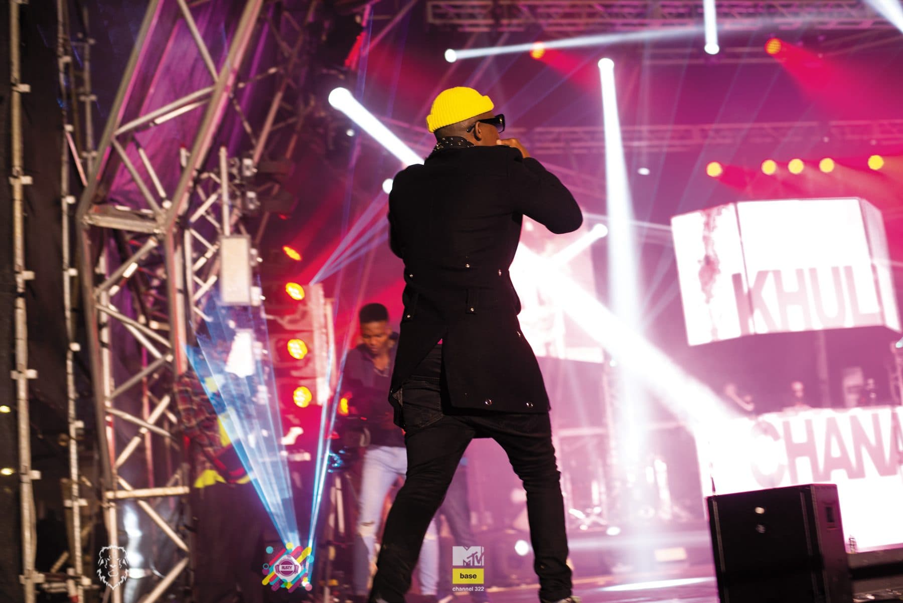 Khuli Chana on stage in a yellow cap under white beams