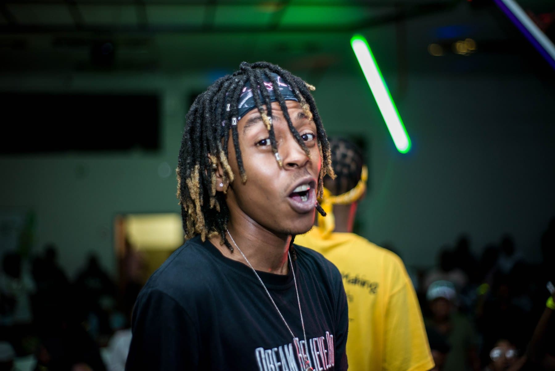Rapper with locs beneath a green light tube
