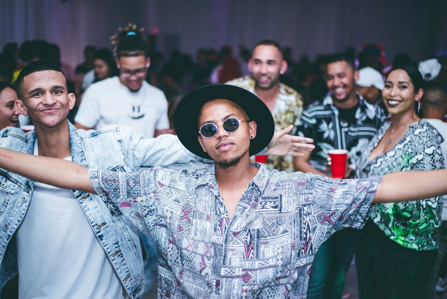 Partygoer in a hat and round glasses, arms wide, friends behind