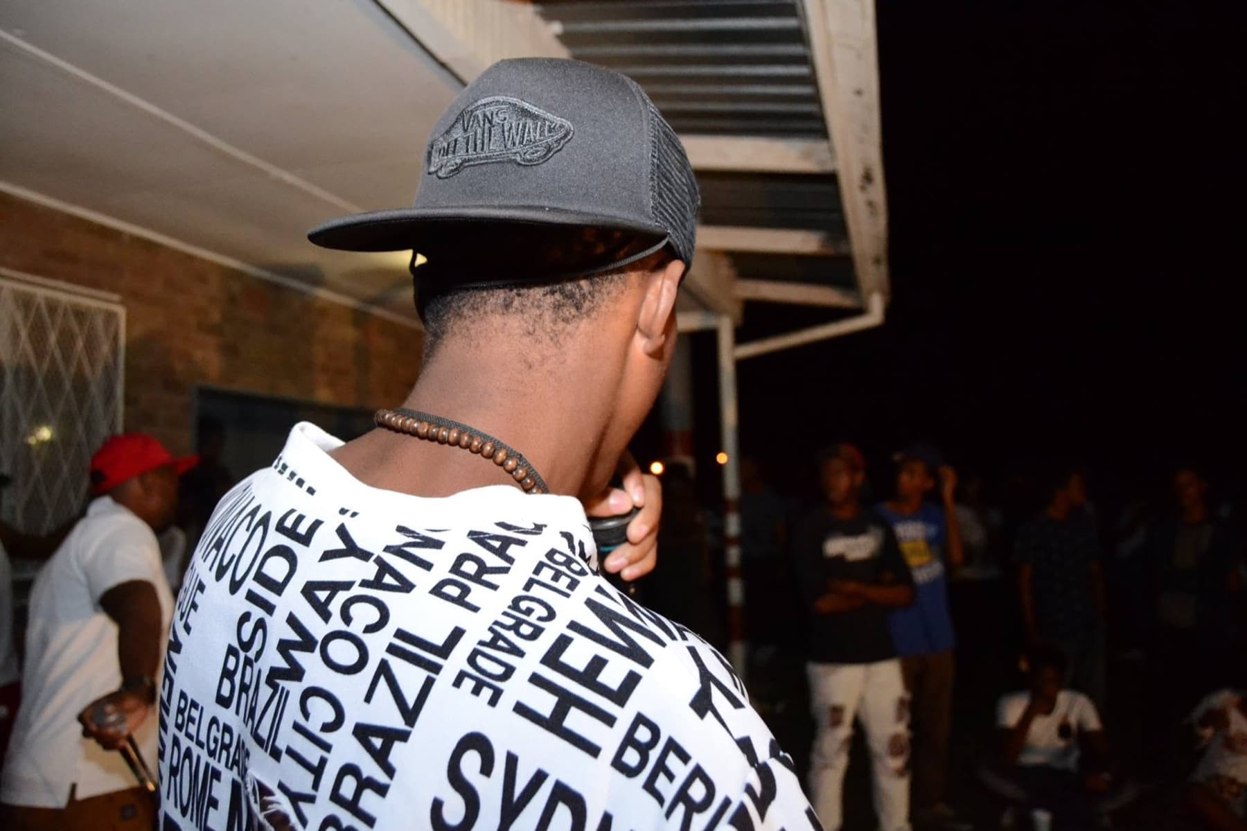 Back of a Vans cap and a city-names shirt, crowd beyond