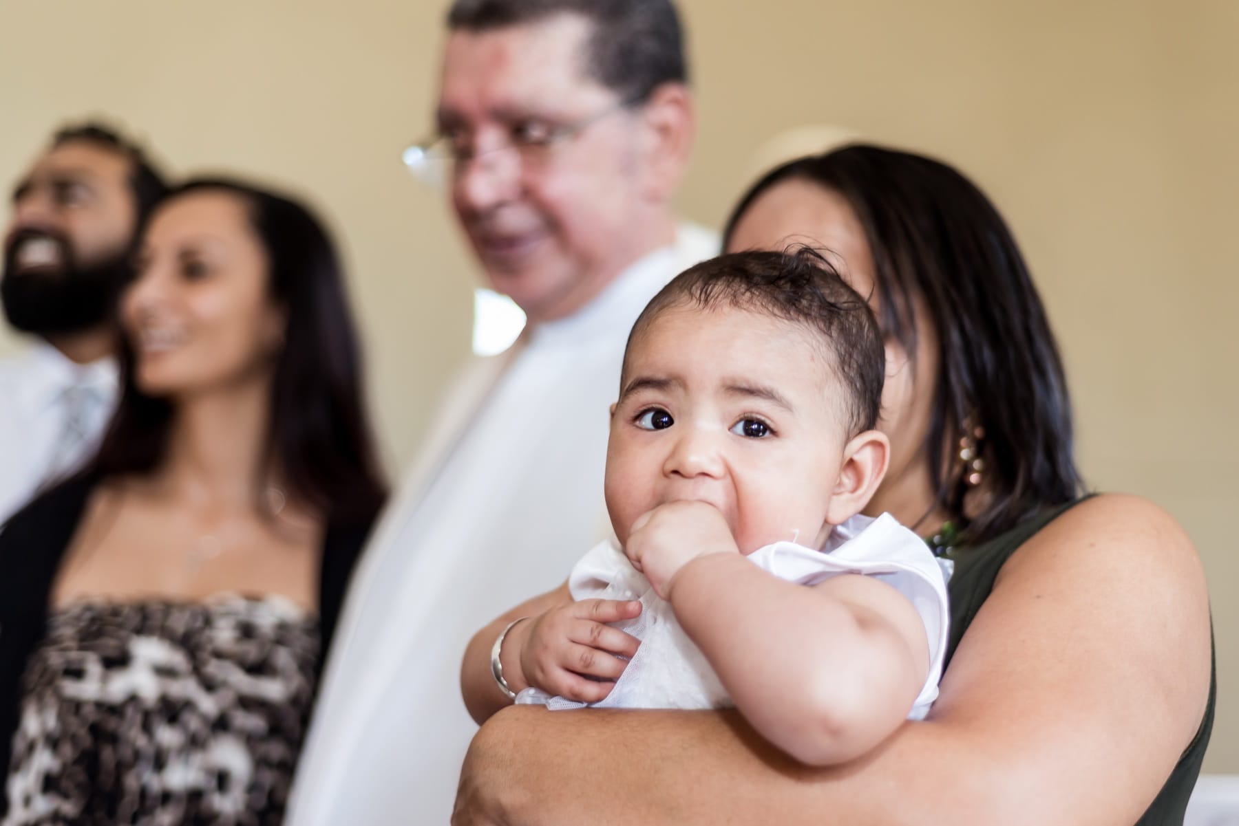 Baby with a fist in her mouth, held through the christening