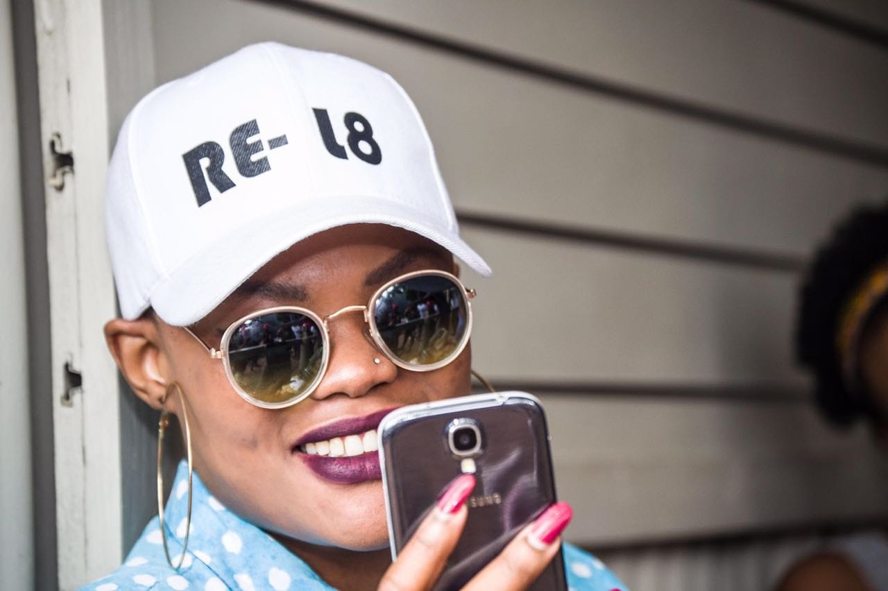 Model in a RE-L8 cap and round shades checking a phone