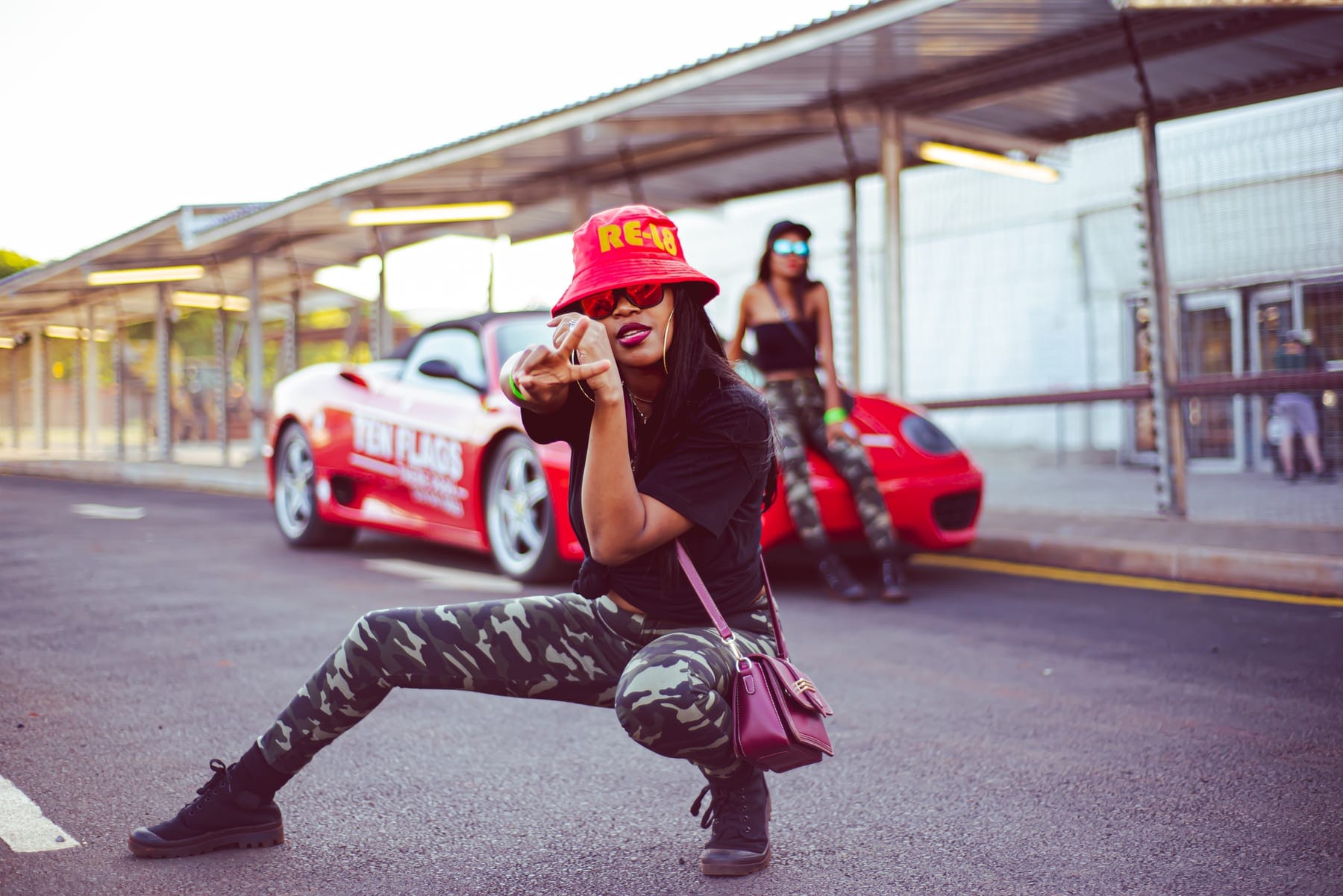 RE-L8 bucket hat and camo, crouched before a red Ferrari