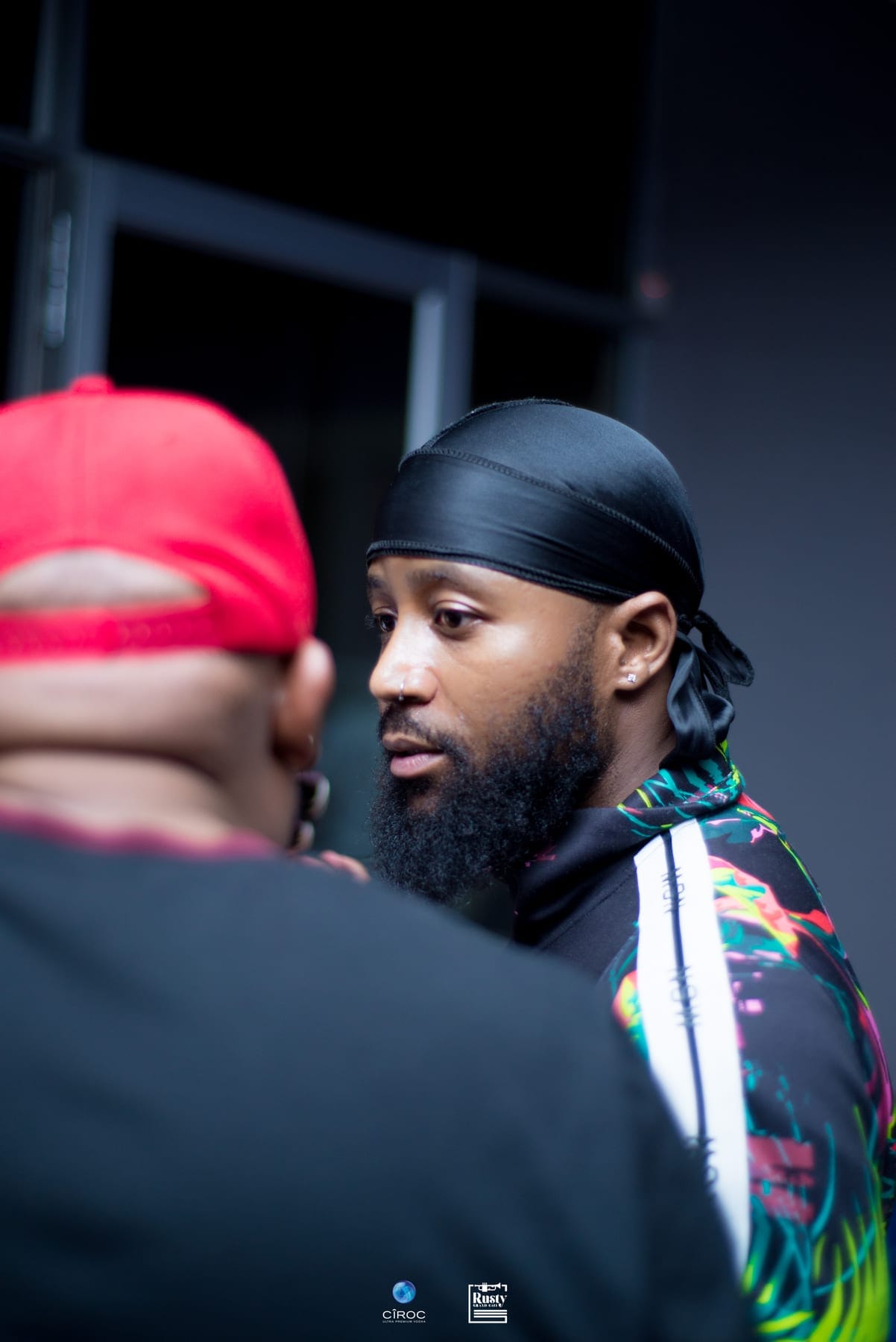 Man in a black durag and a printed track top listening, red cap in the foreground, at Cîroc Nights